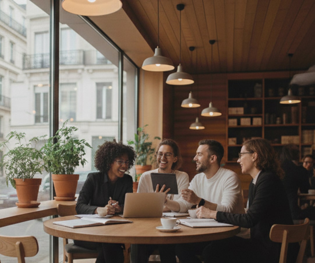 Vier Personen sitzen an einem runden Holztisch in einem Café und diskutieren mit Laptop und Tablet.